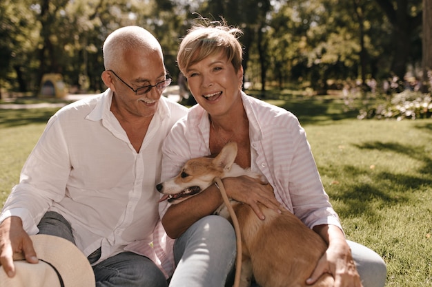 Free photo cheerful lady with short blonde hair in pink shirt laughing, hugging dog and sitting on grass with grey haired man in eyeglasses in park.