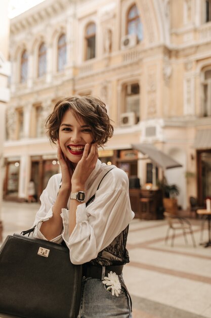 Cheerful lady with wavy hair in blouse with black lace laughing at street. Trendy lady in jeans with handbag posing in city.