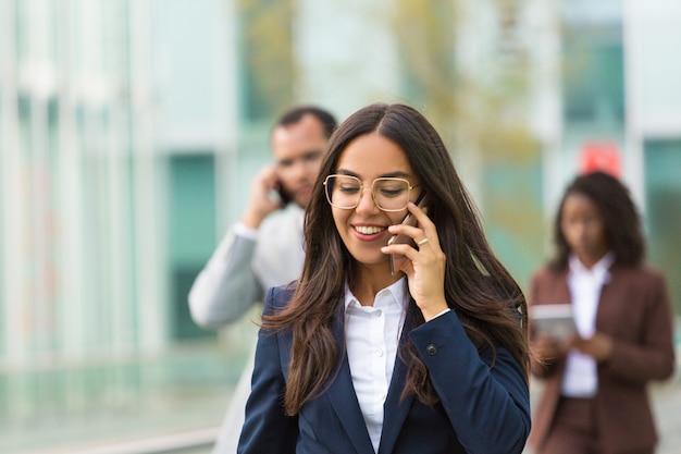 Cheerful Latin businesswoman with phone going down city street