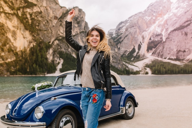 Cheerful laughing girl wearing jeans decorated with embroidery posing near the car on mountain
