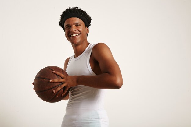 cheerful laughing young African American weaing a white sleeveless shirt and a headband holding a grunge leather basketball to his chest isolated on white.