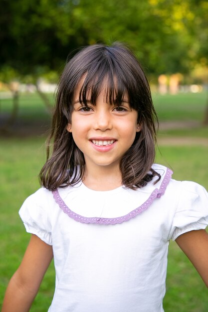 都市公園に立っている陽気な小さな黒い髪の少女。夏の屋外で余暇を楽しむ子供。ミディアムショット、垂直。子供の頃の概念