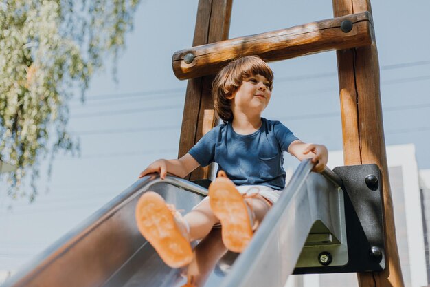 Cheerful little boy in a blue t-shirt and white shorts rolls down a metal slide in city park. child walks outdoors. healthy active recreation. lifestyle. space for text. high quality photo