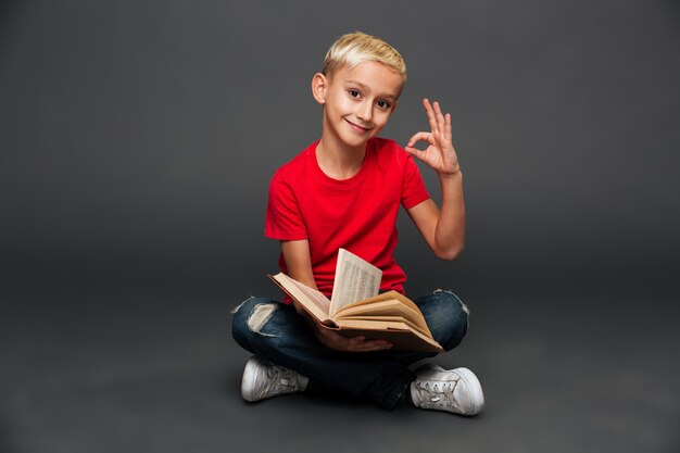 Жизнерадостная книга чтения ребенка мальчика показывая одобренный жест.