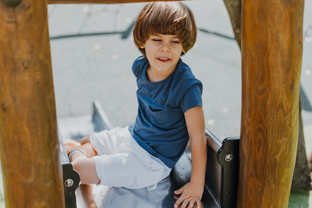Веселый маленький мальчик в синей футболке и белых шортах скатывается с металлической горки в городском парке. ребенок гуляет на свежем воздухе. здоровый активный отдых. образ жизни. место для текста. фото высокого качества