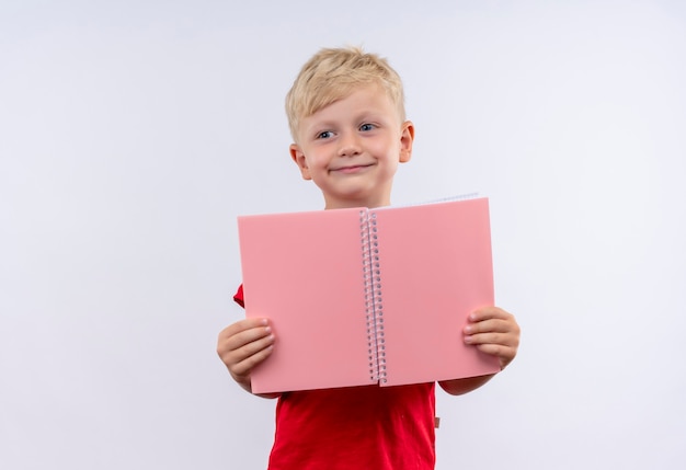 A cheerful little cute blonde boy in red t-shirt showing pink notebook while looking side on a white wall