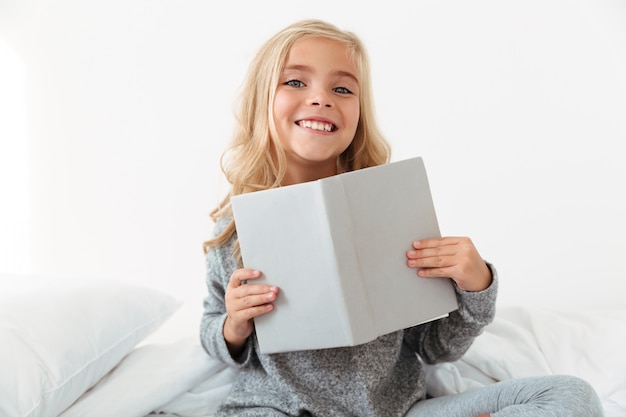 Free photo cheerful little girl in gray pajamas holding book,  while sitting in her bedroom