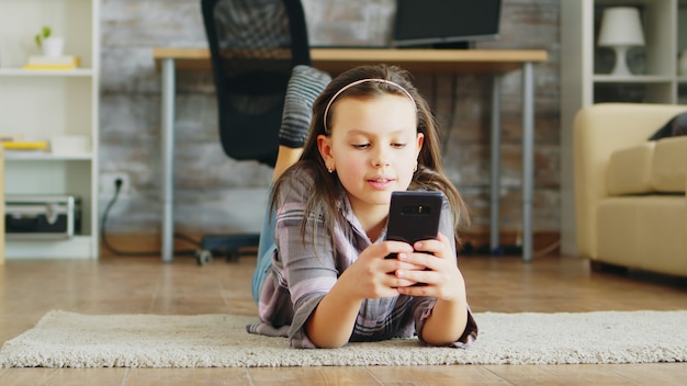 Free photo cheerful little girl lying on the floor carpet using smartphone.