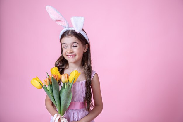 Cheerful little girl with Easter bunny ears smiles and holds a bouquet of tulips in her hands on a pink studio background