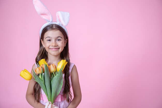 Cheerful little girl with Easter bunny ears smiles and holds a bouquet of tulips in her hands on a pink studio