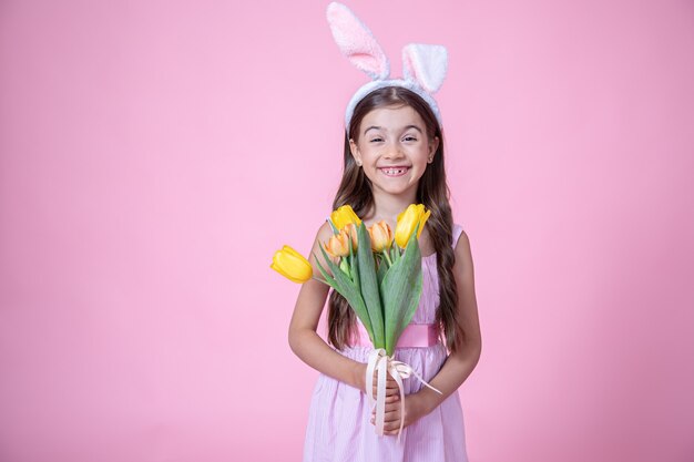 Cheerful little girl with Easter bunny ears smiles and holds a bouquet of tulips in her hands on a pink wall.