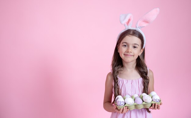 ピンクの壁にイースターバニーの耳と彼女の手に卵のトレイを持つ陽気な少女。イースター休暇のコンセプト。