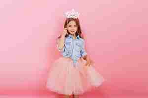 Free photo cheerful little girl with long brunette hair in tulle skirt holding princess crown on head  isolated on pink background. celebrating brightful carnival for kids, birthday party, having fun of cute kid