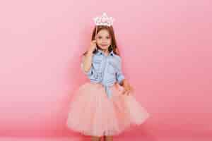 Foto gratuita bambina allegra con capelli lunghi del brunette in gonna di tulle che tiene la corona della principessa sulla testa isolata su fondo rosa. celebrando il carnevale luminoso per i bambini, la festa di compleanno, il divertimento di un bambino carino