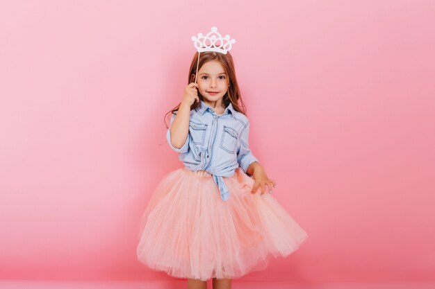ピンクの背景に分離された頭の上の王女の王冠を保持しているチュールスカートで長いブルネットの髪を持つ陽気な少女。子供のための明るいカーニバル、誕生日パーティーを祝って、かわいい子供を楽しんで