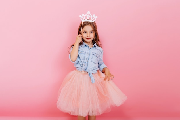 Жизнерадостная маленькая девочка с длинными волосами брюнетки в юбке тюля держит корону принцессы на голове, изолированной на розовом фоне. Празднование яркого детского карнавала, дня рождения, веселья милого малыша