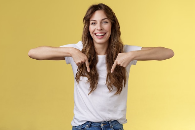 Cheerful lively happy funny girl long beautiful hair pointing down index fingers indicate bottom promo laughing joyfully share positive emotions recommend product advertising yellow background