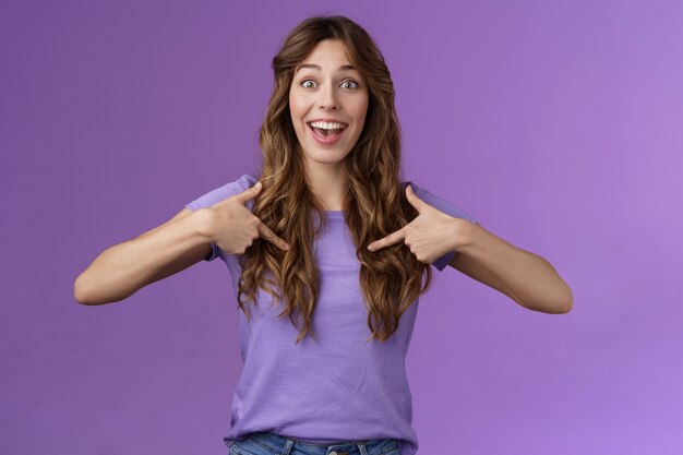 Cheerful lively upbeat entertained happy girl curly hairstyle open mouth amused pointing center herself happily tell own achievement smiling broadly telling friends promotion purple background