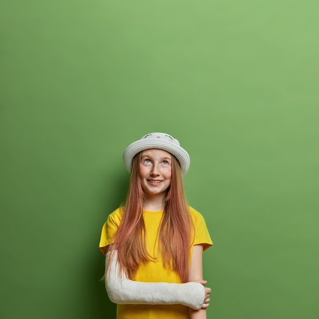 Cheerful long haired teenage girl with broken arm in plaster cast after reckless bicycle driving, wears summer hat and yellow t shirt, looks happily above, hopes for fast recovery. Children, accidents