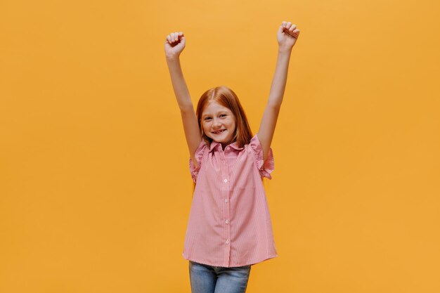 Cheerful longhaired redhead little girl in jeans and striped shirt rises arms and rejoices on isolated orange background