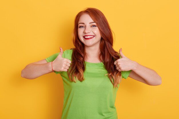 Cheerful lovely girl showing thumbs up while posing isolated, red haired girl in green t shirt