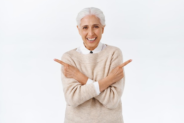 Cheerful, lovely senior woman with grey combed hair, wear fancy elegant clothes, pointing hands sideways with crossed arms over chest, smiling and laughing