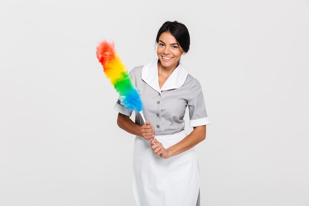 Cheerful maid in uniform holding colorful duster while standing