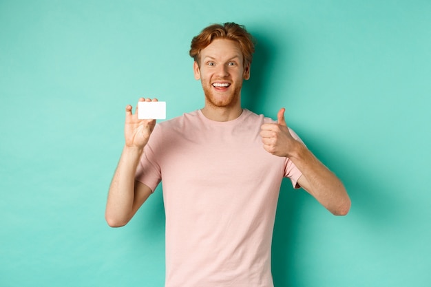 Cheerful male bank client in t-shirt showing thumb up and plastic credit card, smiling satisfied at camera, standing over turquoise background.
