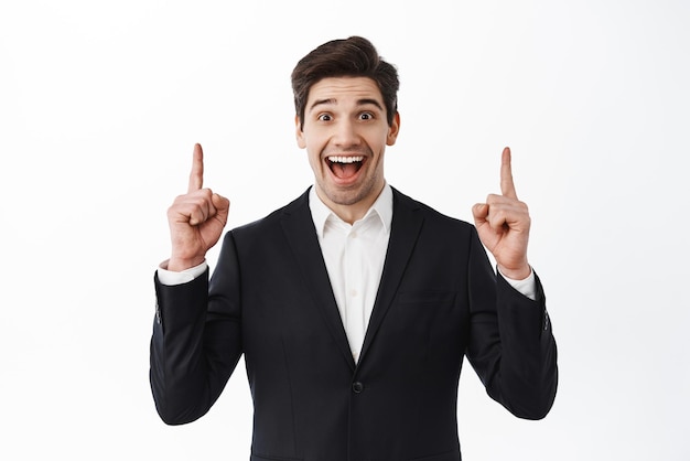 Cheerful male estate agent broker in suit pointing fingers up showing amazing offer best promo sale smiling amused standing over white background