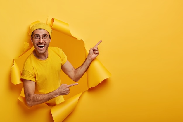 Free photo cheerful male gives nice offer, advertises new product on sale, stands in torn paper hole, has positive expression