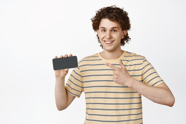 携帯電話の画面に指を指し、白い背景の上にオンラインで立っているアプリストアを示す笑顔の陽気な男性モデル