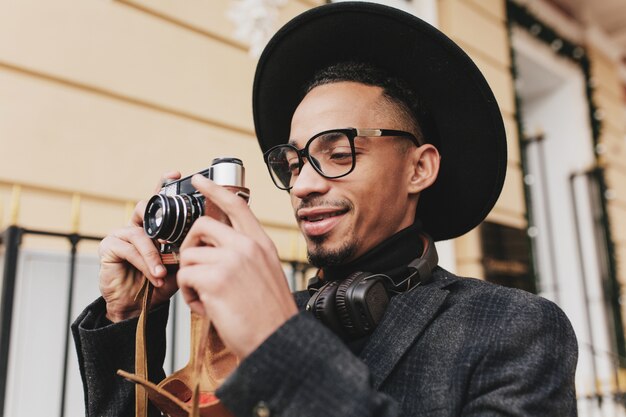 朝の屋外で働く茶色の肌を持つ陽気な男性写真家。ポジティブなアフリカ人の写真は、カメラを持って通りに立っている暗い服を着ています。