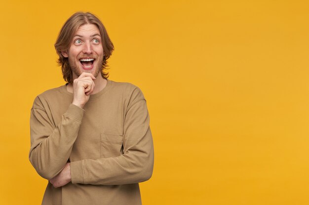 Cheerful male, positive and excited bearded guy with blond hairstyle. wearing beige sweater. touching his chin and smiling. watching to the right at copy space, isolated over yellow wall