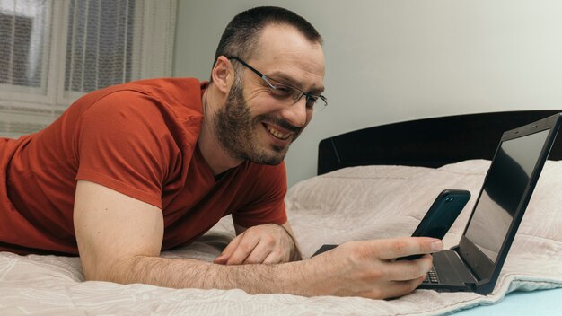 Cheerful man browsing laptop and smartphone on bed