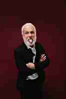 Free photo cheerful man demonstrates tongue on burgundy background funny guy with beard in black suit and light shirt posing on isolated backdrop