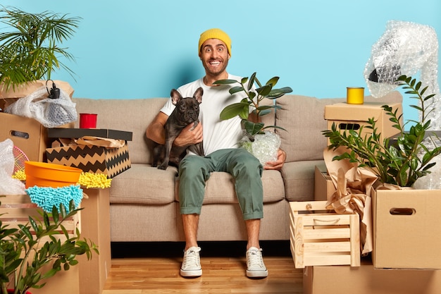 Cheerful man embraces dog and pot with houseplant, sits in living room on sofa