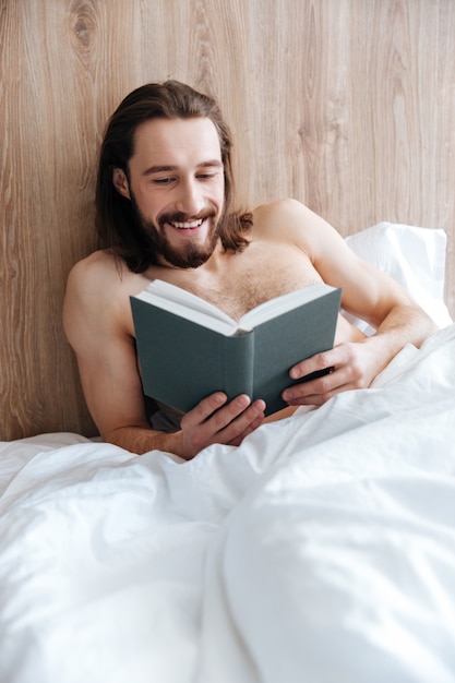Cheerful man lying and reading book in bed