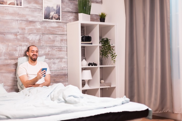 快適なベッドで夜にスマートフォンで面白いビデオを見ながら笑っているパジャマの陽気な男。