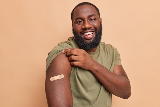 Cheerful man shows adhesive plaster on shoulder after getting coronavirus vaccine feels safe gets injection in arm cares about health during pandemic isolated over beige wall