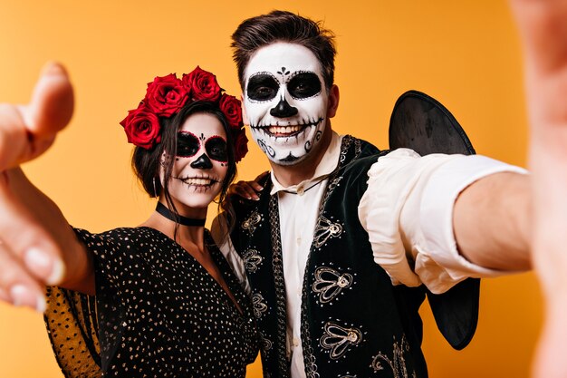 Cheerful man in sombrero making selfie with girlfriend in halloween. Funny guys with zombie makeup chilling on orange wall.