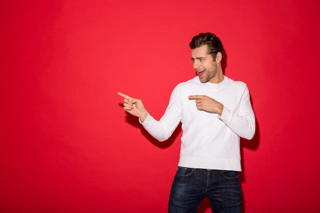 Cheerful man in sweater pointing and looking away with open mouth over red wall