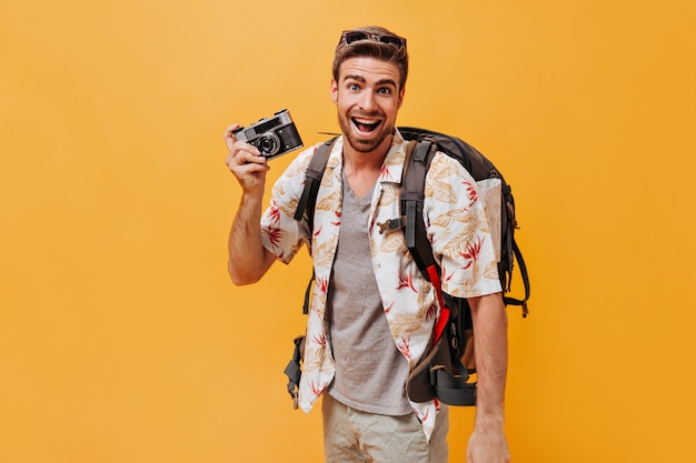 Cheerful man with beard in grey t-shirt and printed light shirt smiling and posing with camera and backpack on orange wall