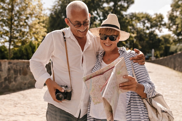 Cheerful man with grey hair in light shirt and jeans with camera smiling and looking at map with blonde lady in hat and blue outfit in park.