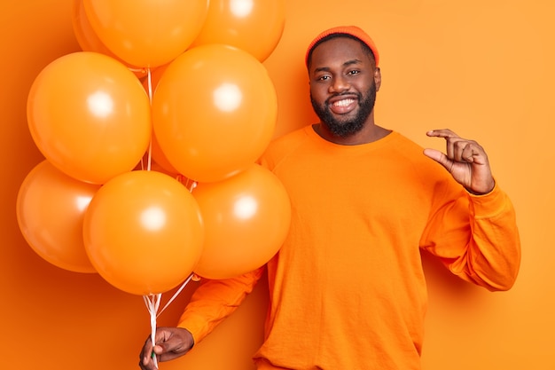 Free photo cheerful man with inflated balloons makes tiny gesture says he needs not much time to prepare for party dressed in casual clothes isolated over vivid orange wall