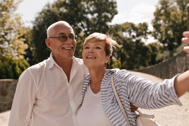 Cheerful man with mustache in glasses and light shirt smiling with woman with short hair in striped blue clothes in park.