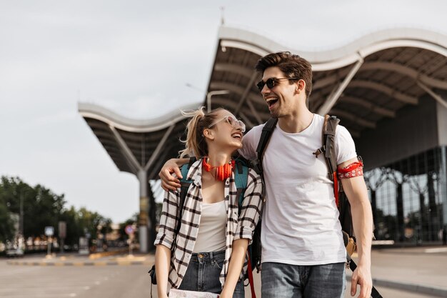 サングラスをかけた陽気な男と女が空港の近くで笑う白いTシャツを着た男と女が地図とバックパックを持っています