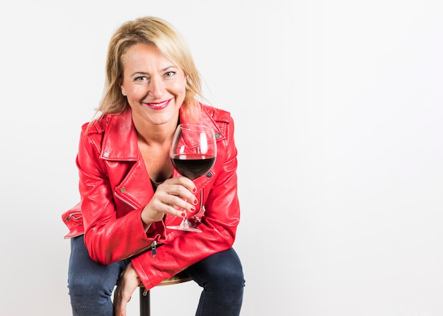 Cheerful mature woman sitting on stool holding wine glass in hand isolated on white background