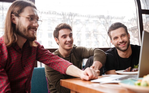 Cheerful men friends sitting in cafe while eating. Using laptop.