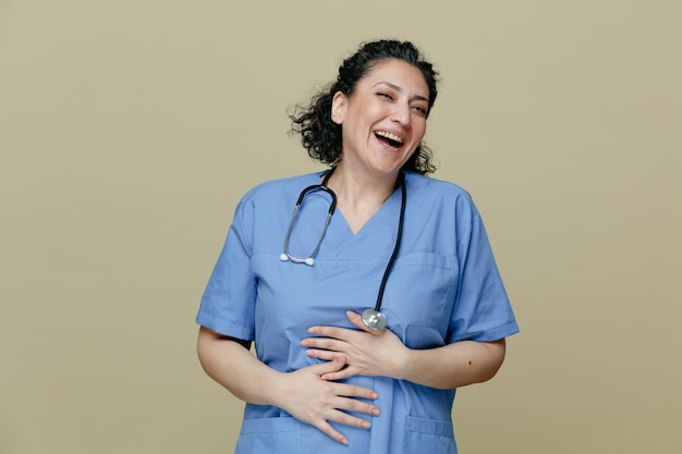 Cheerful middleaged female doctor wearing uniform and stethoscope around neck keeping hands on belly looking at camera laughing isolated on olive background