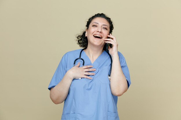 쾌활한 중년 여성 의사는 제복을 입고 목에 청진기를 두르고 올리브 녹색 배경에 격리된 가슴에 손을 얹고 웃으면서 전화 통화를 하는 쪽을 바라보고 있습니다.